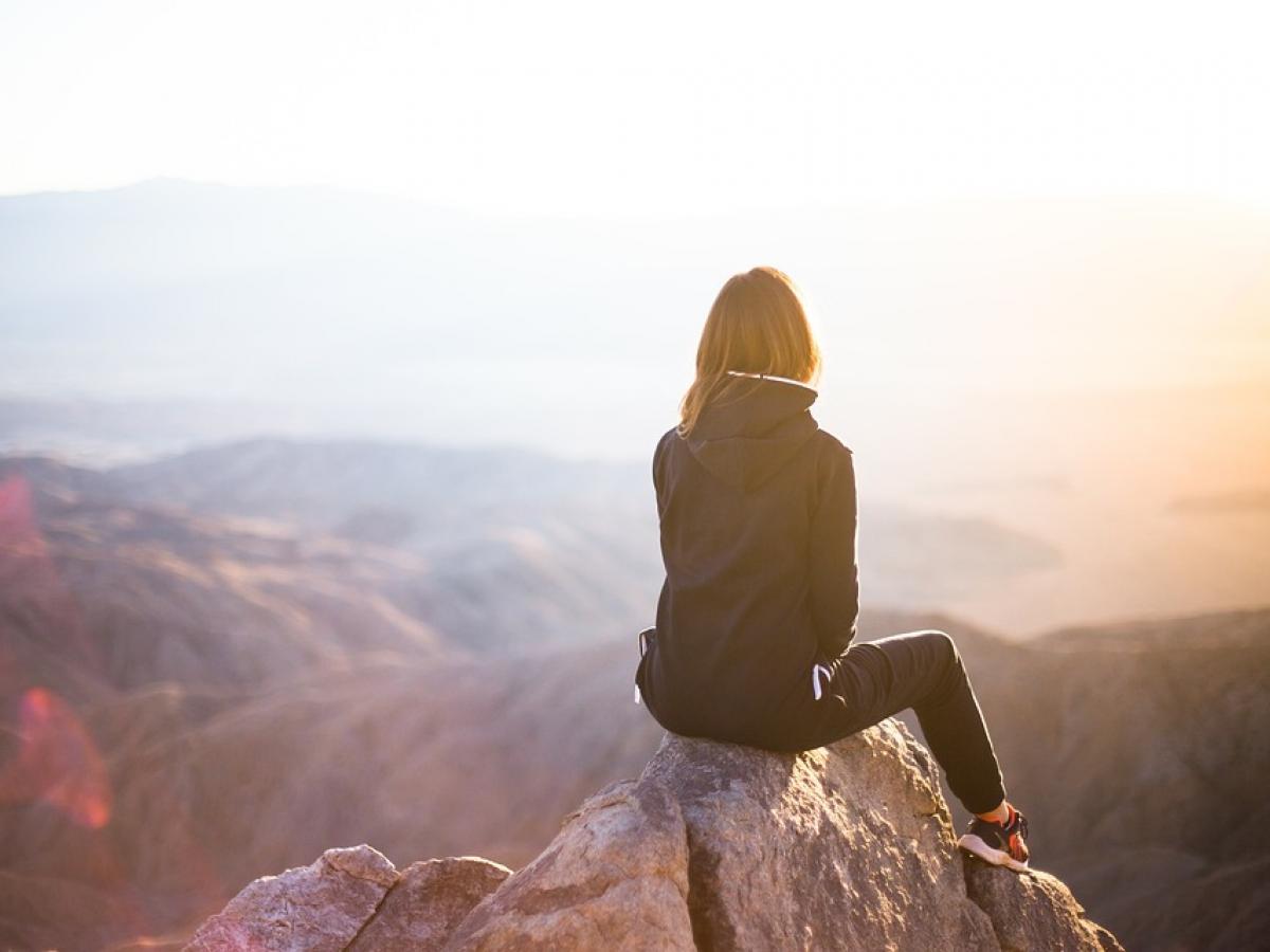 A woman sitting on a boulder staring over mountains and valleys at sunset.