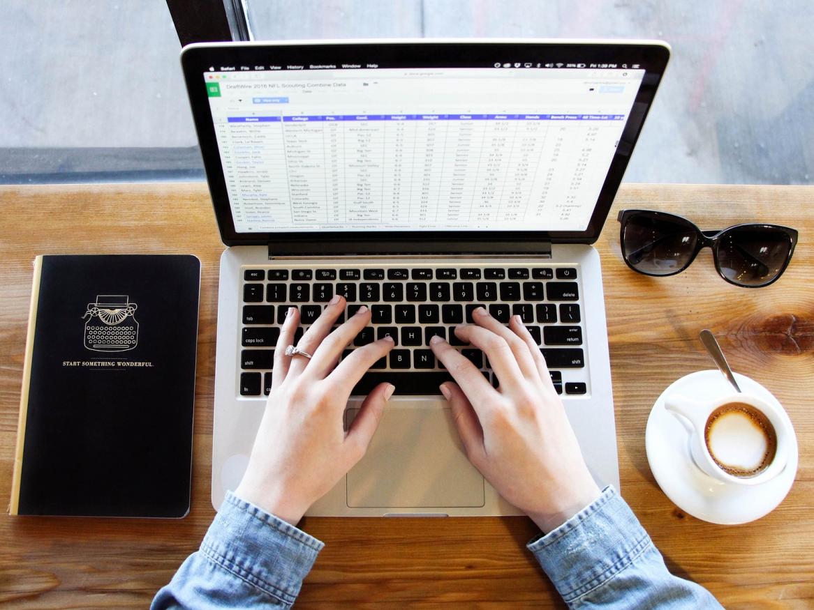 A laptop, coffee cup, sunglasses and notebook on a wooden table with hands hovering over the keyboard