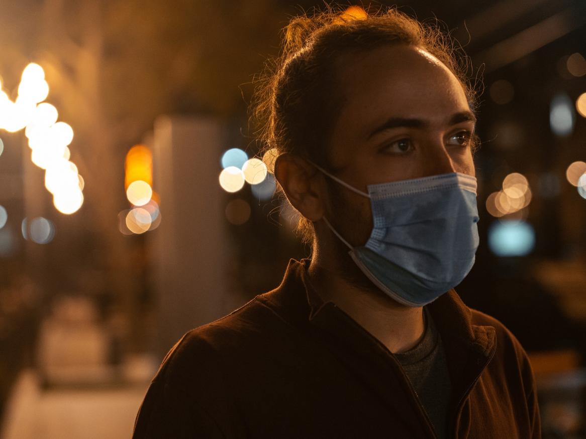 Person wearing a COVID mask walking down a darkened street with lights in the background