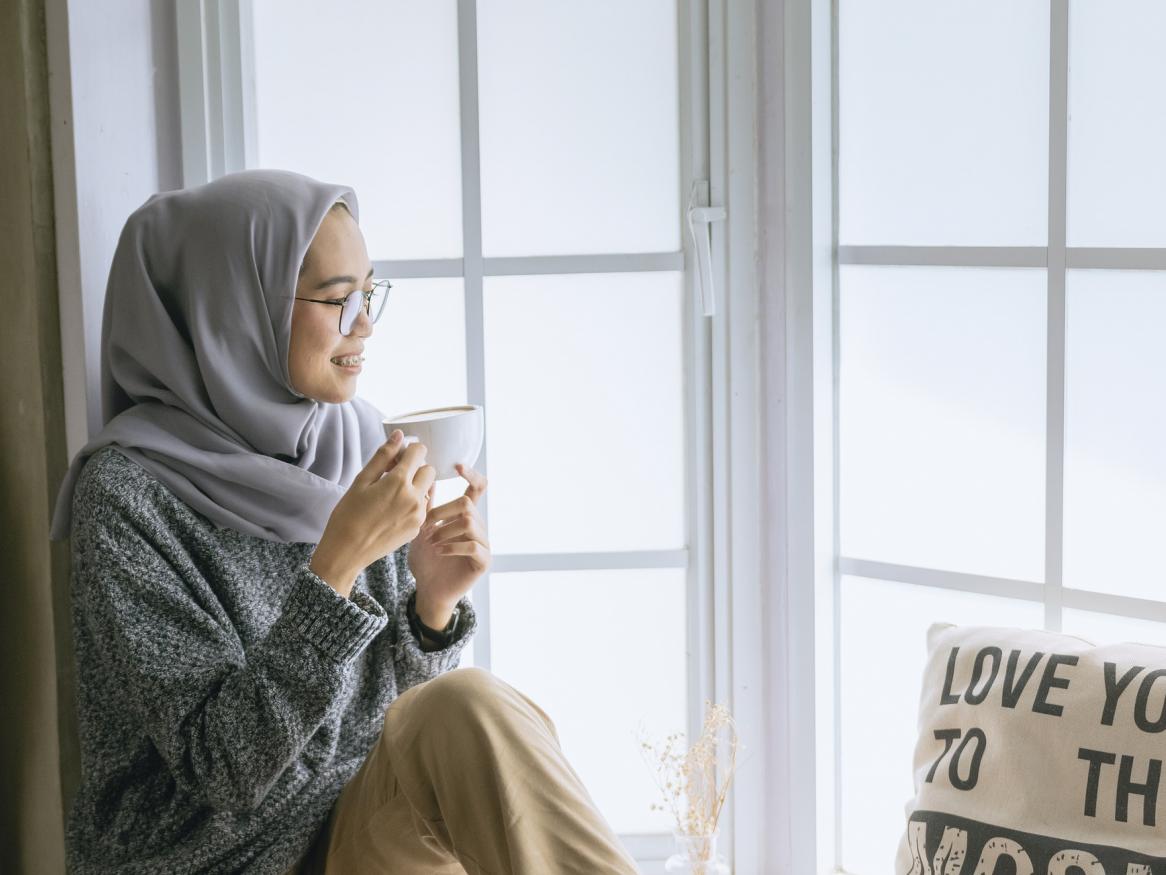 Women sitting in a window sill drinking a cup of tea and looking out the window smiling