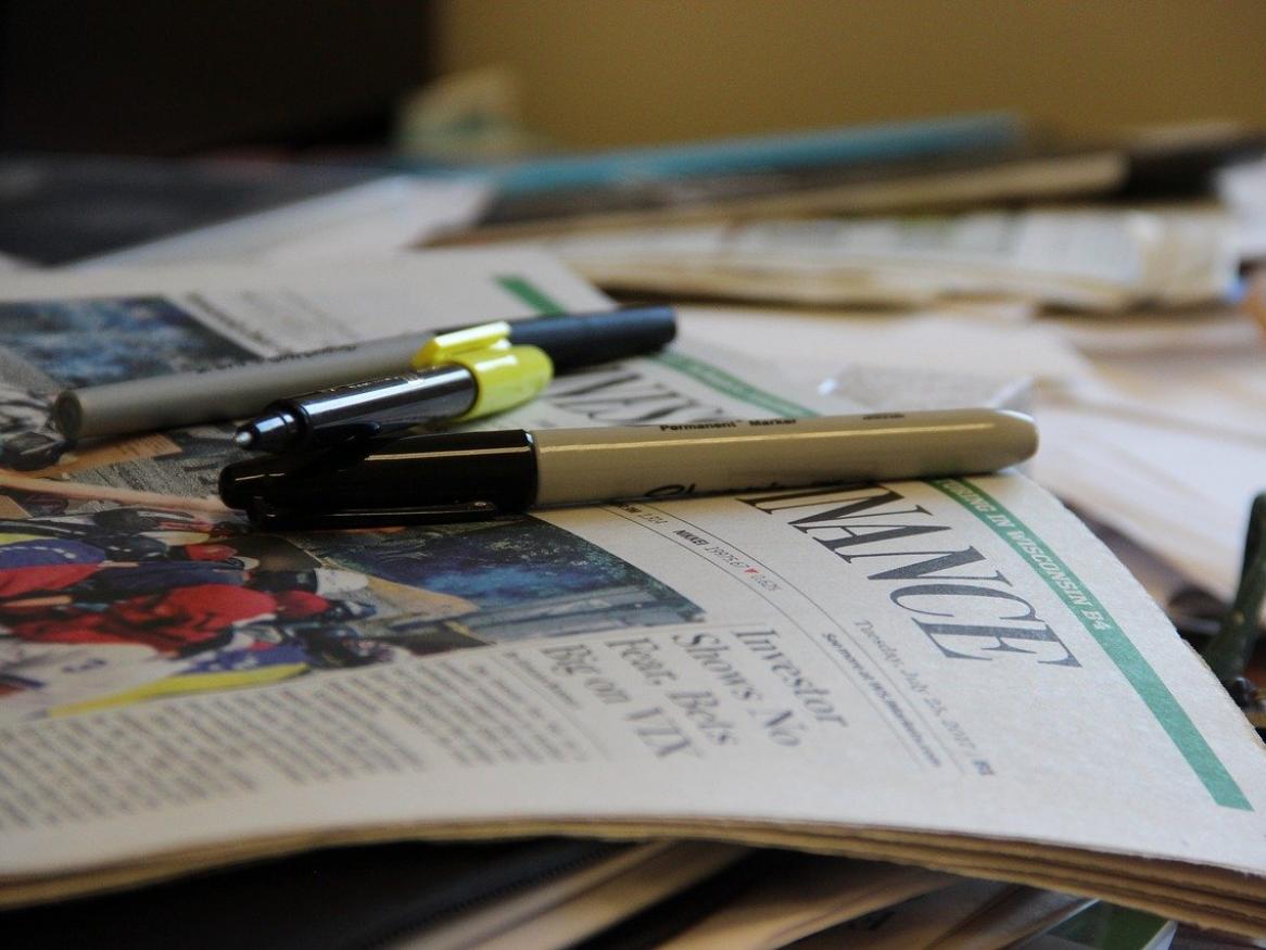 An image of a messy desk, with pens and highlighters on top of a newspaper and stacks of paper