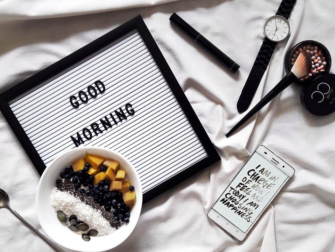 An image of a sheet with a sign reading 'GOOD MORNING', a bowl of fruit, a watch, a phone, and makeup brushes.