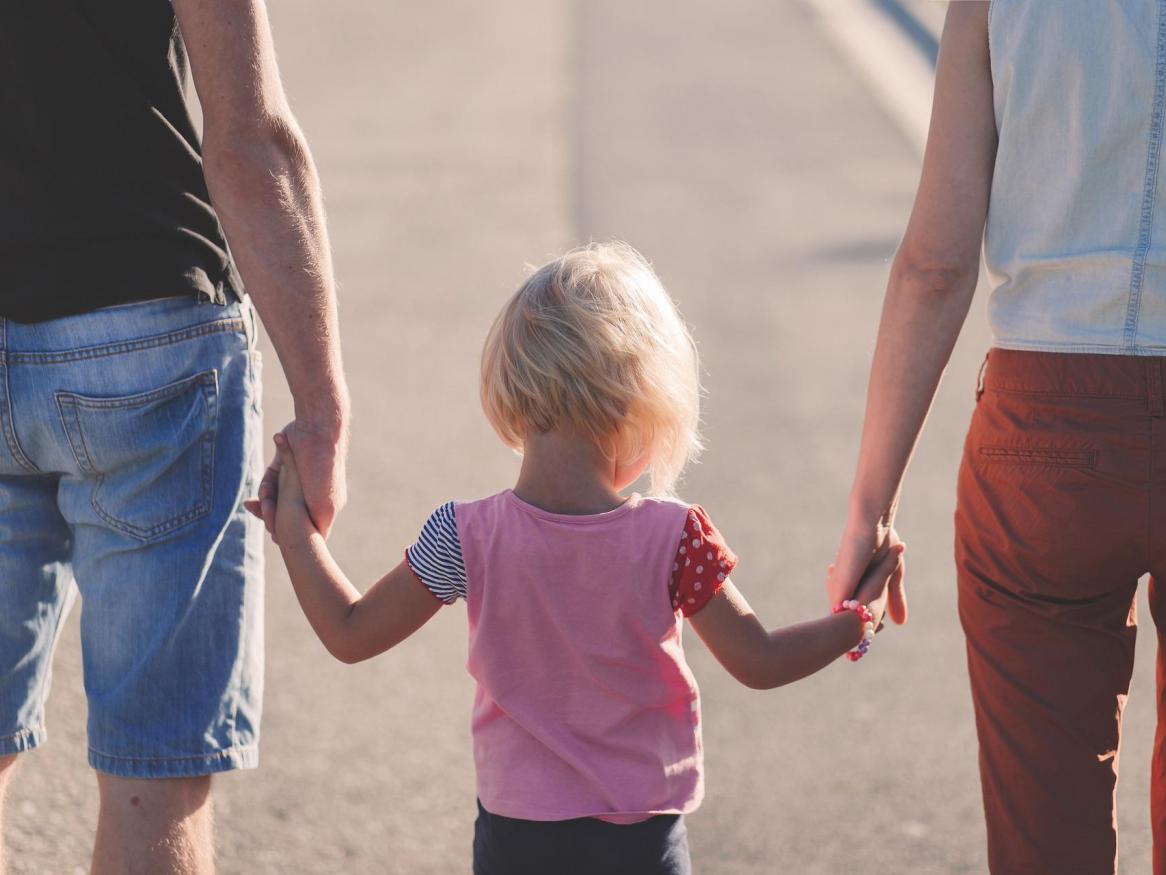Rear body shots of 2 adults holding either hand of a toddler, walking away from the camera.