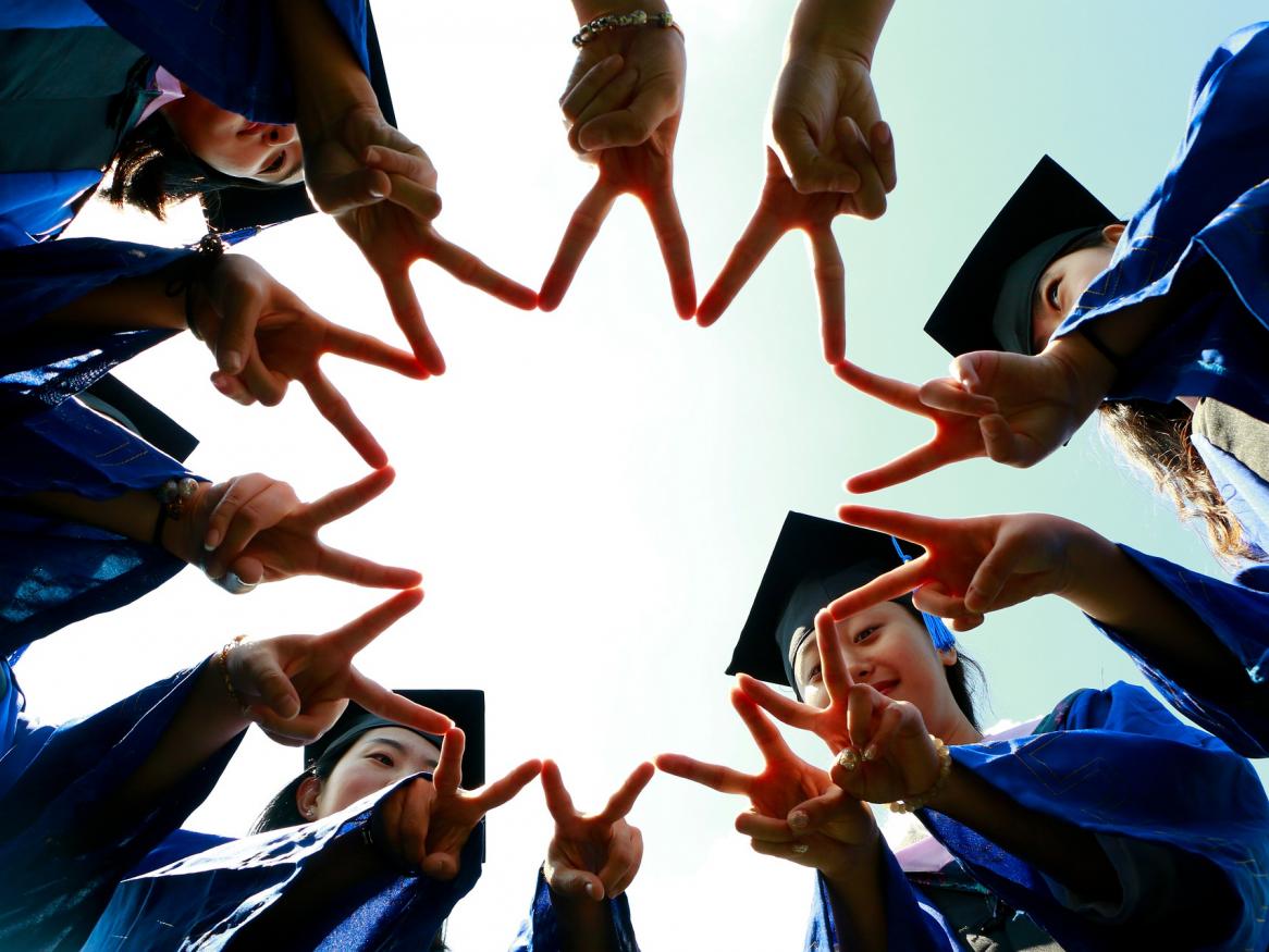 view from below of 6 students in graduation gowns holding their fingers in v signs together in a group