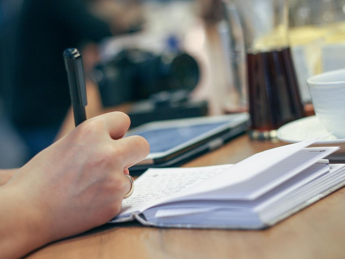 Photo of a hand with a pen, writing on a pad on a desk