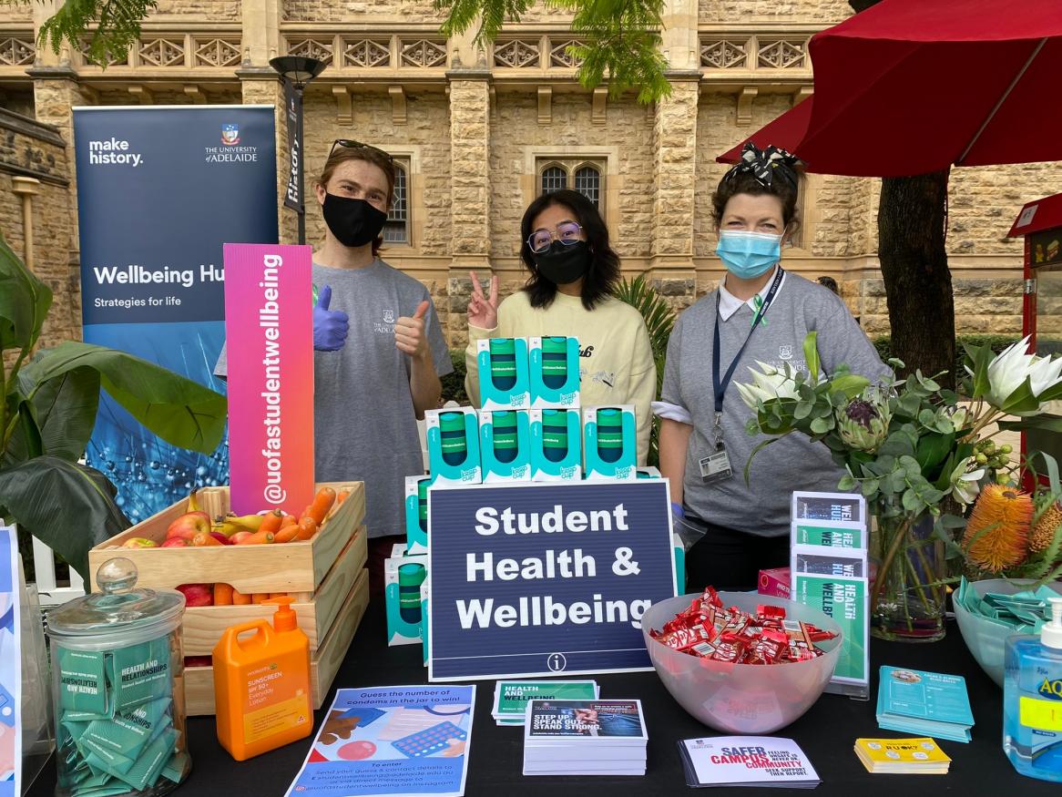 Photo of 3 people wearing masks standing behind a table with giveaways including keep cups, condoms and postcards
