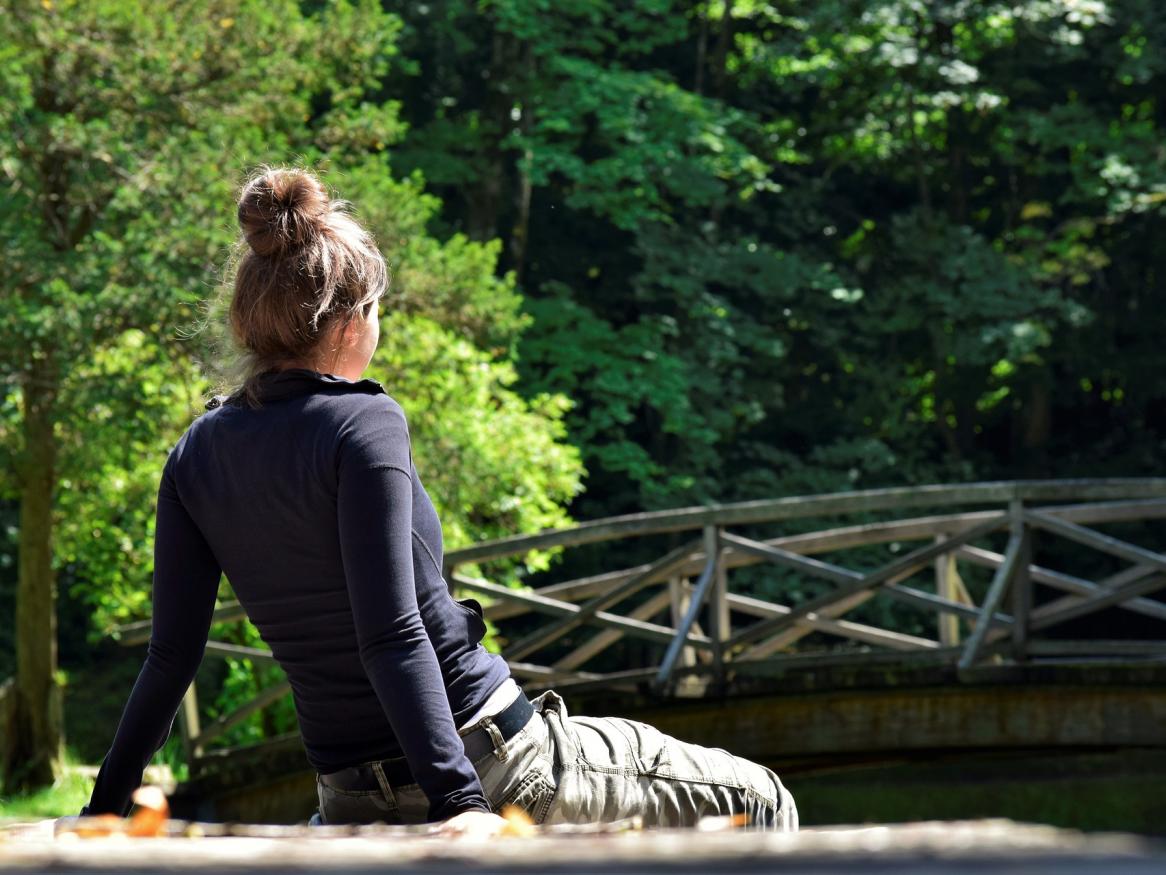 photo of the back of a girl sitting looking at a bridge in a forest landscape