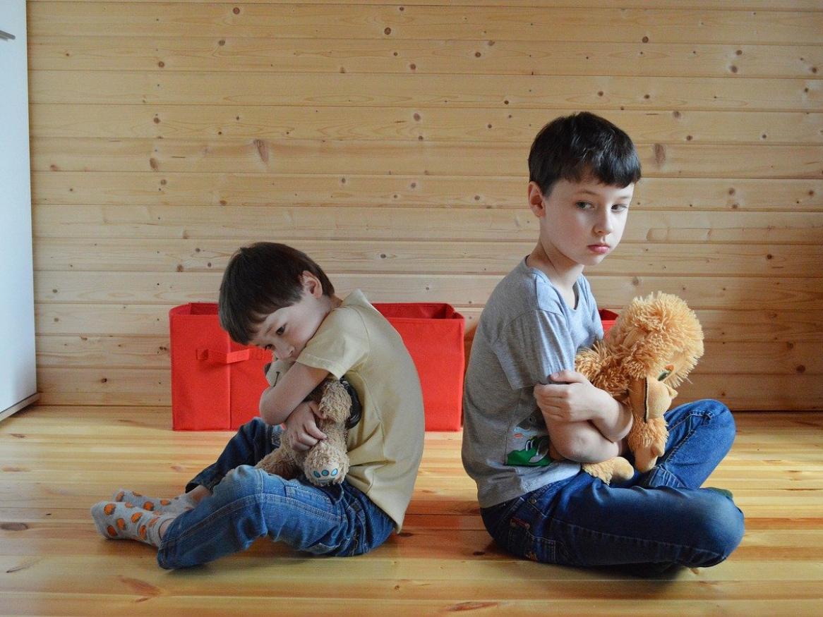 Two kids sit crossed legged, back to back, holding a teddy bear each. 
