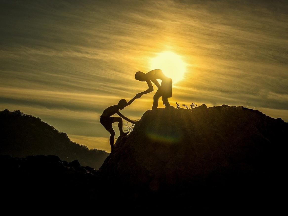 A person helps another person climb up a hill, with the sunset behind them.