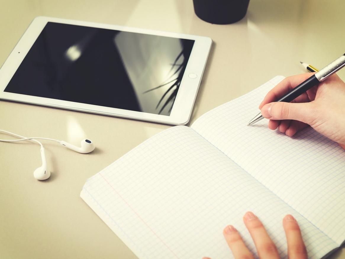A person begins to write in a blank notebook on a table with their iPad and headphones.