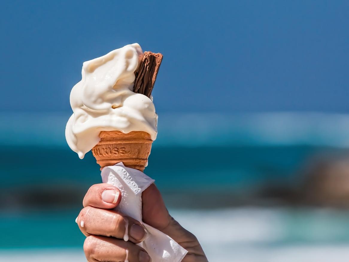 A hand holding a vanilla ice cream cone with a chocolate stick, in front of a blue ocean.