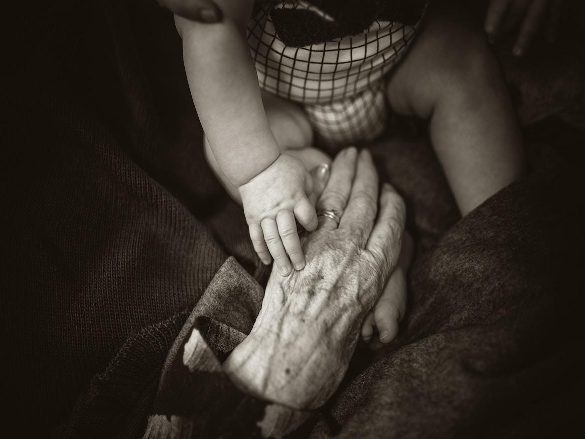 An elderly person holding hands with an infant.