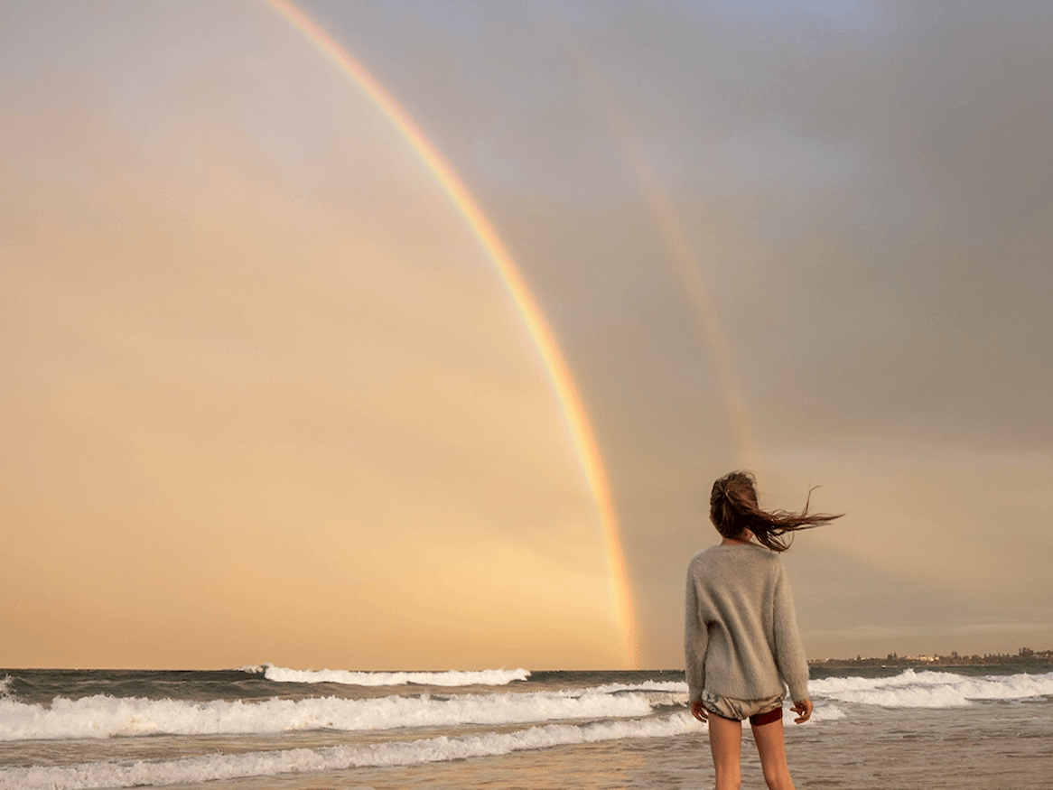 A girl facing a rainbow while standing on the beach