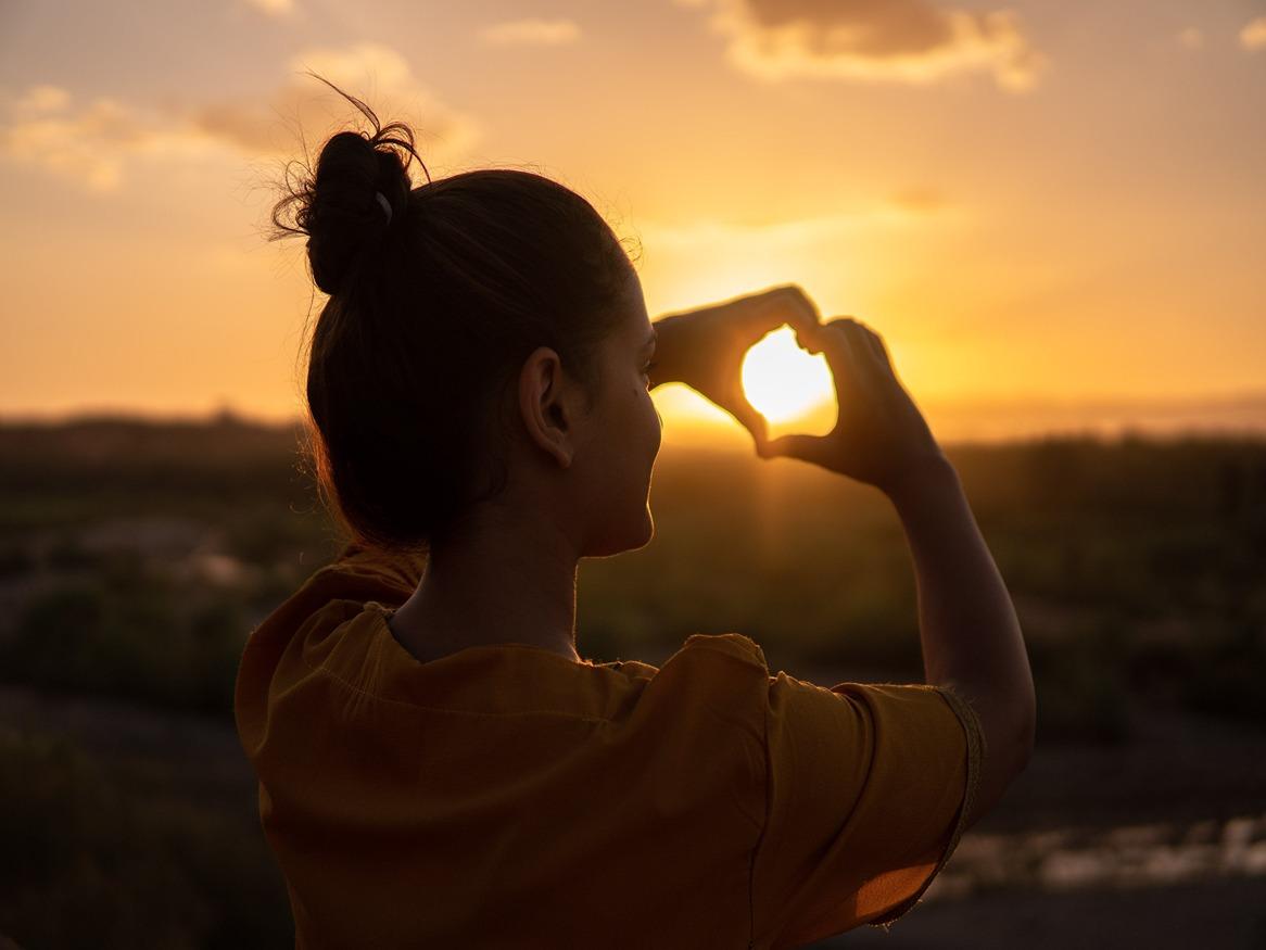 A woman creating a heart with her hands in front of a sunset