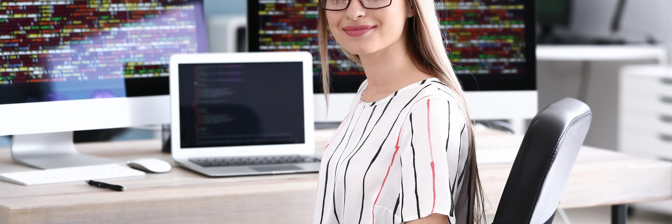 A young woman sits at a desk by a laptop with coding screens in background